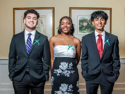 Three young people in formal wear, two boys and one girl, smile at the camera in a ballroom.